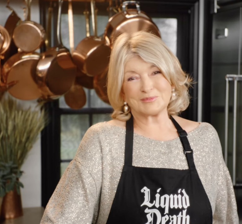 A smiling Martha Stewart in a kitchen wearing a black Liquid Death apron over a beige sweater.