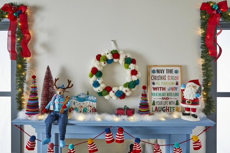 A blue holiday mantle, with a garland of red and white striped hats. A red, white, green and teal puff ball garland hangs on the wall over the mantle, and a variety of decorative trees, a Santa figurine, a reindeer figurine holding skis and wearing a ski outfit, a holiday sign, a ski themed advent calendar and faux snow and lights rest on top of the mantle.