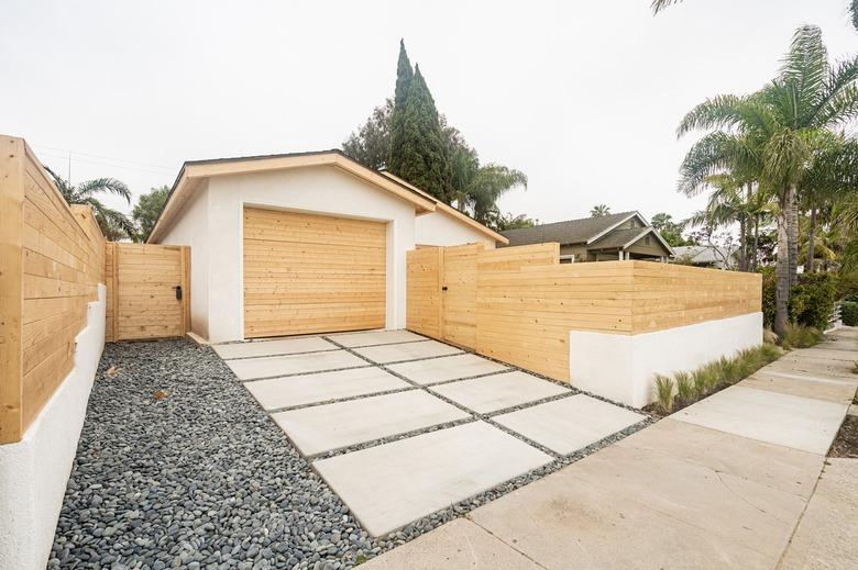 Concrete and gravel walkway leading to a white garage with a wood door surrounded by a wood wall.