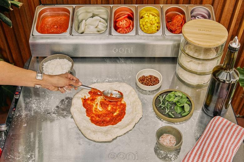 A person spreading tomato sauce on top of pizza dough on a countertop with containers of toppings