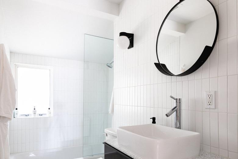 A minimalist all white tiled bathroom with a round black framed mirror