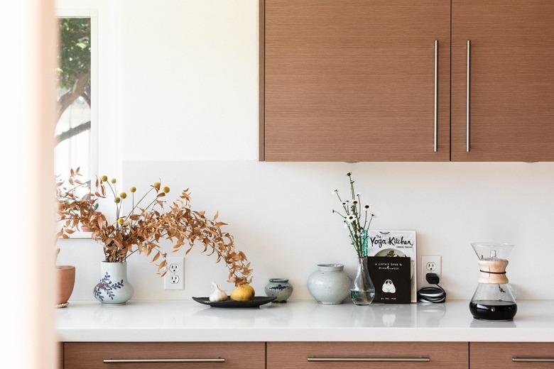 Wood kitchen cabinets, and a white counter with vases of flowers, pottery pieces, and a Chemex coffee.