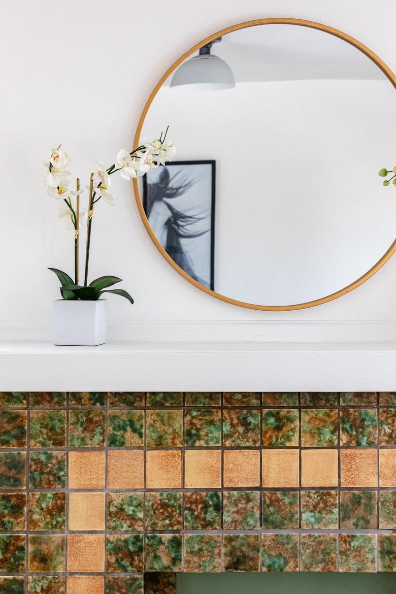 A decorative fireplace with colorful tiles, and large round mirror and white orchids