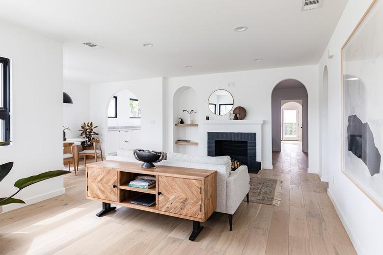 Contemporary living room with hardwood floor, white walls, grey couch, black fireplace, plants, and arched hallway