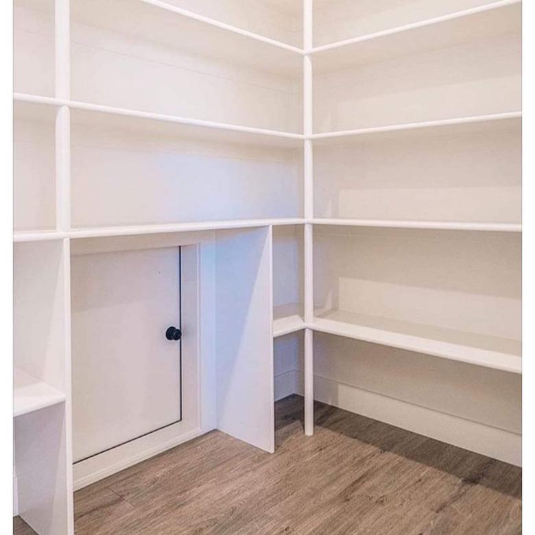 A white pantry with shelving above a small square door with a black doorknob.