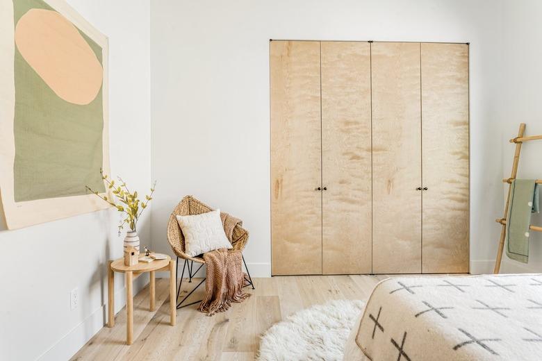 Bi-folding closet, accent chair with white pillow, neutral throw, wood stool with vase of flowers, abstract wall hanging