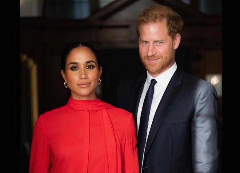 Meghan Markle in a red dress and Prince Harry in a black suit standing next to each other.