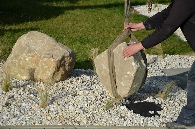 safe handling of a very heavy granite boulder with a crane, the stone is picked up and tied with straps and placed in a hole a gloved worker stabilizes the stone to the flowerbed