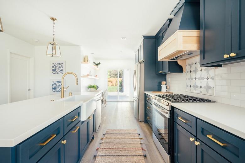 Dark blue kitchen cabinets with gold handles and white countertops. Ornate backsplash by stovetop. Gold lantern pendant lights.