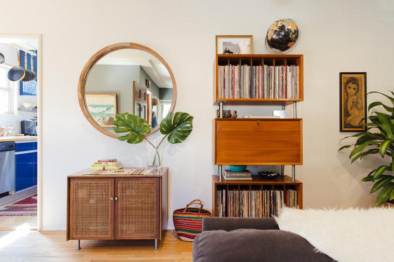 Wall featuring midcentury shelving unit displaying vinyl records, wood circular mirror, and small midcentury wood and rattan accent table