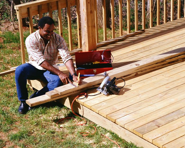 A carpenter sits on a deck measuring wood to complete a construction project.