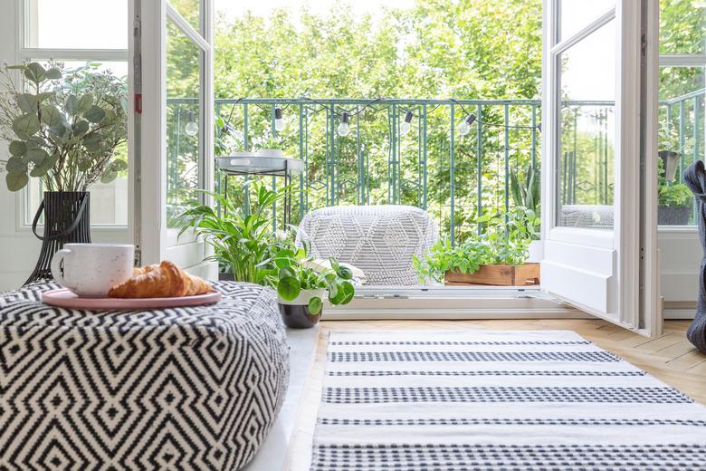 Pouf with pink plate with coffee mug and croissant in blurred foreground in real photo of room with fresh plants, rug on the floor and open door to balcony