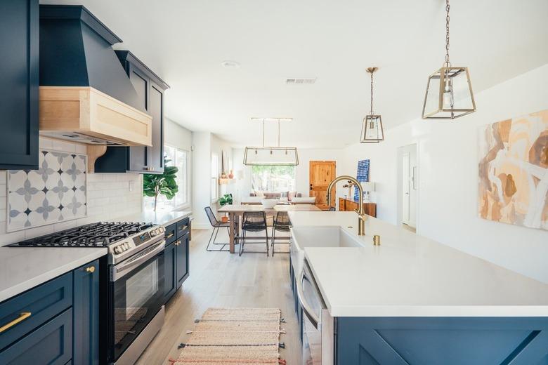 Dark blue kitchen cabinets with gold handles and white countertops. Ornate backsplash by stovetop. Gold lantern pendant lights.