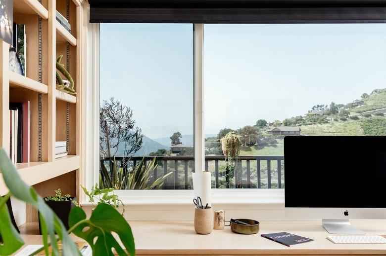 A light wood desk with a computer and containers. A bookcase is beside. A window view overlooking hills and neighboring homes.