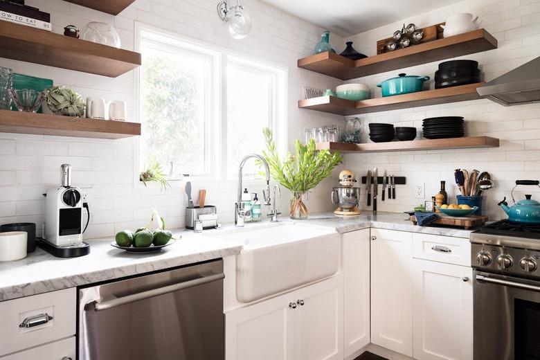 A kitchen corner with white cabinets, a stainless steel dishwasher, oven, and open wood shelving stacked with black, white, and blue servingware.