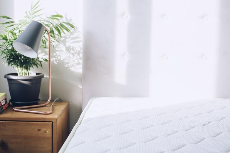 White mattress beside side table with small plant and lamp against white curtains.