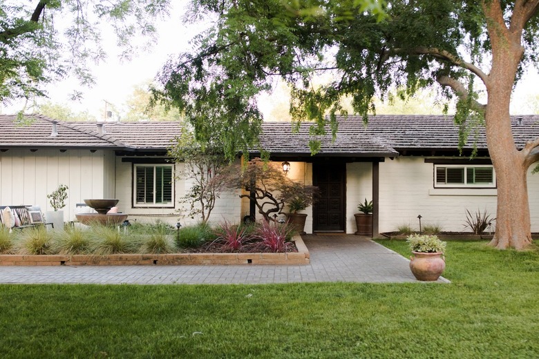 Ranch style white home with a large tree and a flowerbed with a wood retaining wall in the front