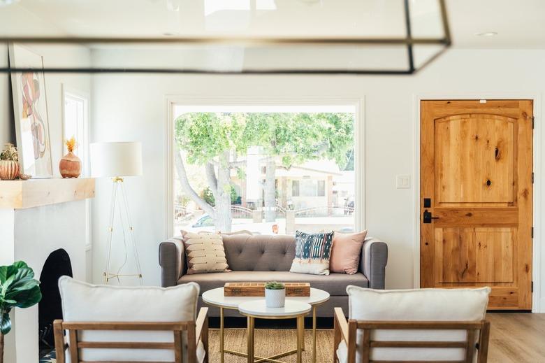 A living room with wood door, gray couch with throw pillows and white side chairs.