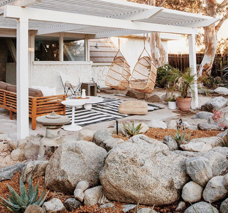 White wood modern pergola with hanging chairs, couch, outdoor rug and rock garden.