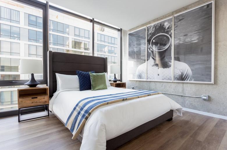 Modern loft bedroom with wood headboard, large photo on wall, wood floors, wood nightstand, black lamp, white lampshade, large industrial windows.