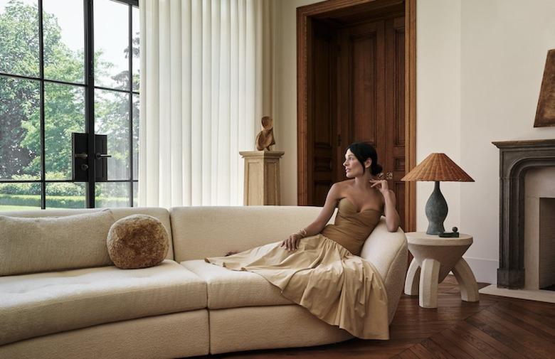 Athena Calderone in a beige top with a lighter skirt laying on a beige couch looking out a large window