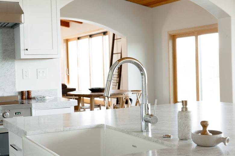 In a kitchen, a sink in a marble countertop, part of a kitchen island. The chrome faucet is running water. Next to the sink, a marble soap dispenser and a wooden scrub brush in a ceramic dish.