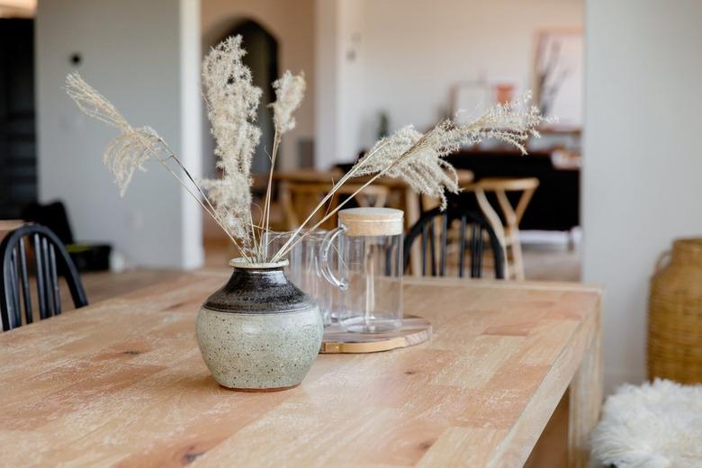 Light wood tabletop with a grey and black ceramic vase with stalks of wispy grass in it. Behind the vase, there