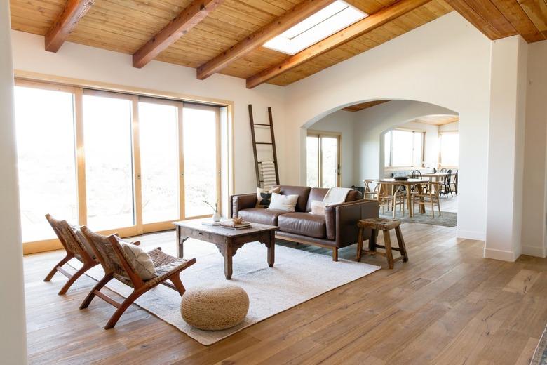 A large, open living room with white walls, sliding glass doors, and a high ceiling with wooden beams. There is a skylight in the ceiling. In the center of the room, a rustic wooden coffee table on a white rug. On the far side, there