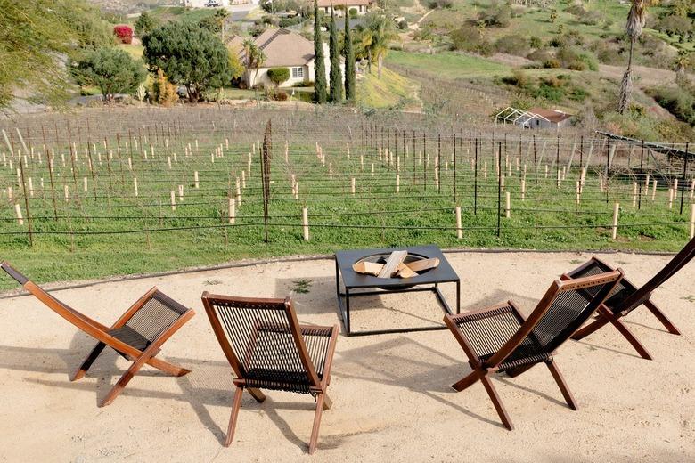 Dirt patio area with four wooden and rope lounge chairs and a fire pit. The seating area overlooks a young vineyard.