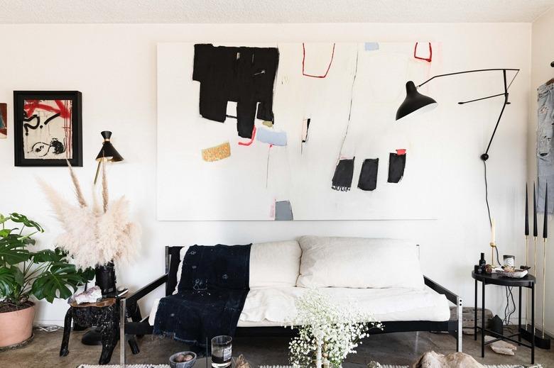 Abstract art over a white cushion sofa. Black lamps and side tables on each side. Houseplant and dried florals for decor.