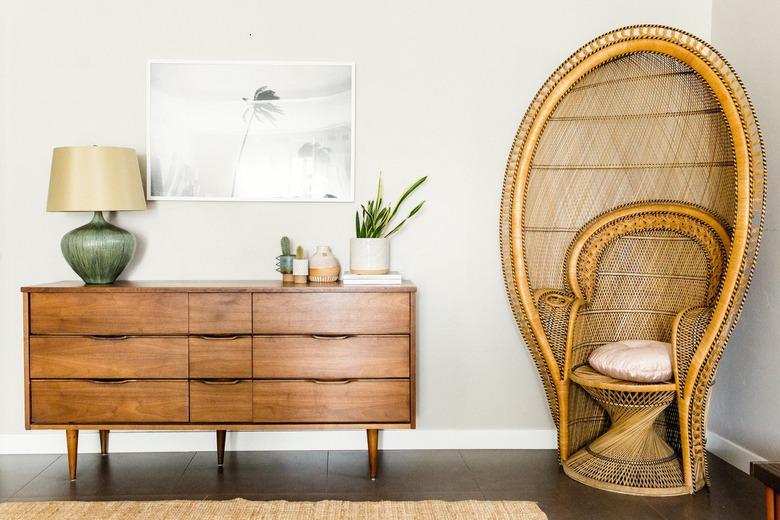 A peacock chair with a round cushion, next to a wood sideboard with plants and a vintage lamp. A wall photo of palm trees.