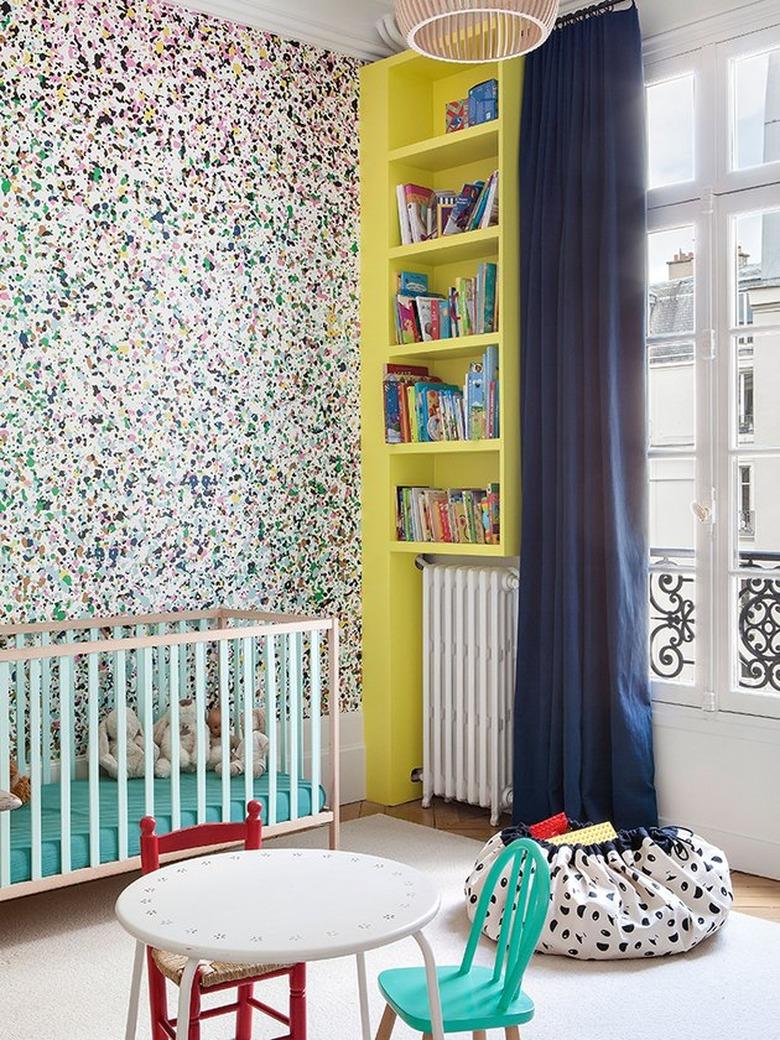 Modern nursery with paint splatter pattern wallpaper, yellow built-in shelves, modern crib, bean bag.