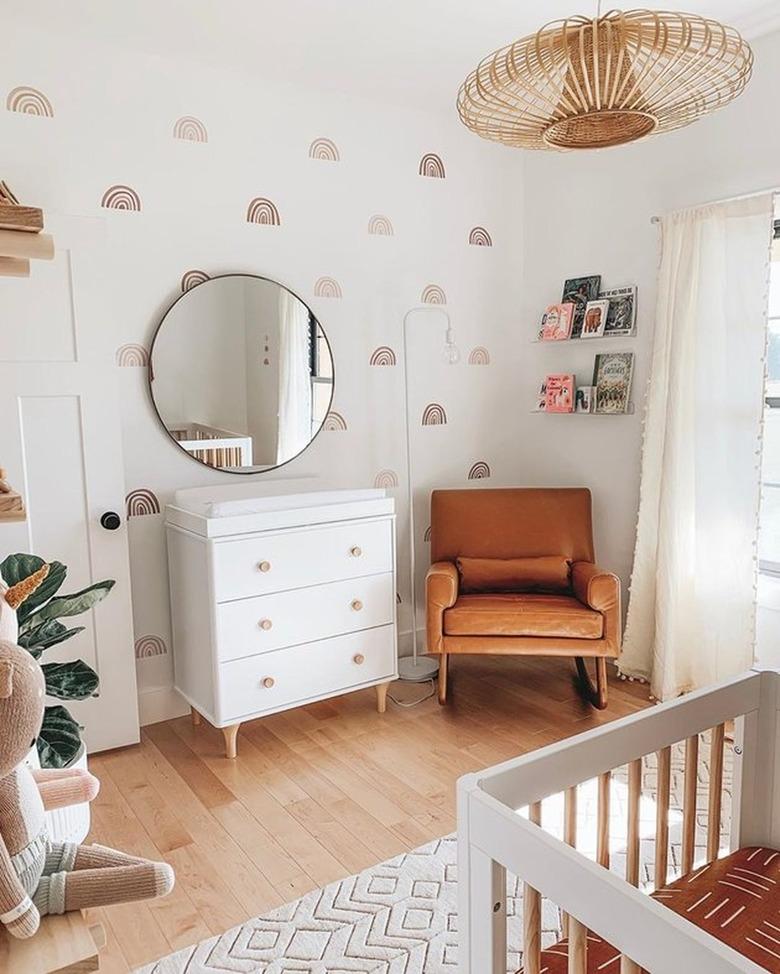 Modern nursery with rainbow pattern wallpaper, white dresser, chair, cane lamp.