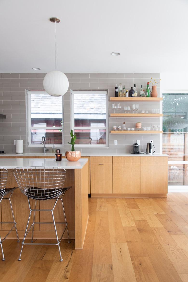 Kitchen with wood cabinets, gray wall tiles, round white pendant light, wall shelves, and Modernist metal wire stools.