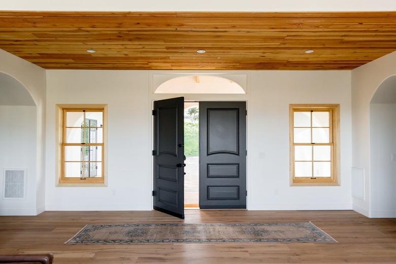 An entry hall of a Spanish-style home with double dark wood doors, one of which is open, on a white wall. Two windows with light wood frames on either side of the door. The floor and ceiling are both made of wood. There's a rug in front of the door.