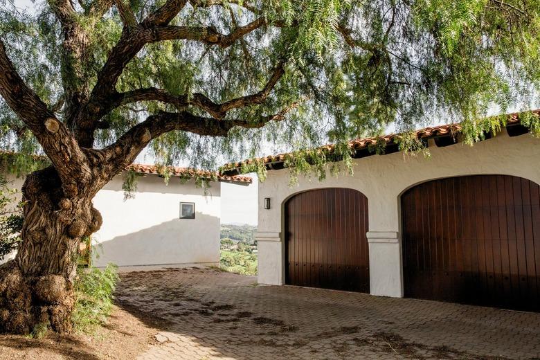 Brick driveway leading to a white Spanish-style garage with two wood doors. Oak tree by a driveway.