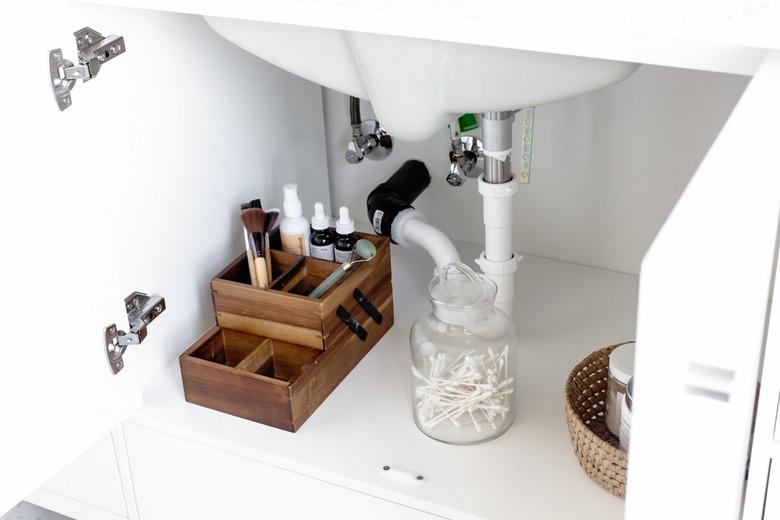 Plumbing of a sink in a vanity. Beside there is a wood organizer with makeup and a jade roller. A glass container with q-tips is beside.