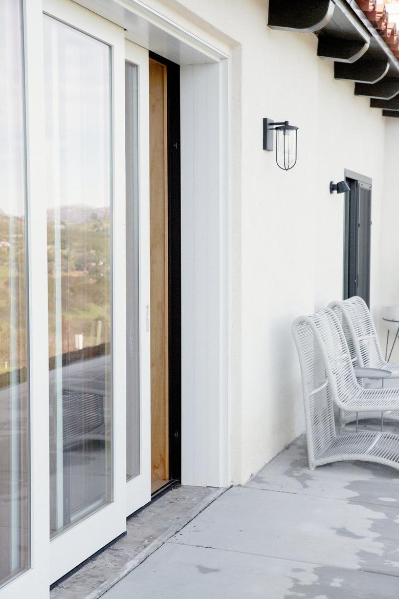 White wall with a glass sliding door, partially open. A black metal sconce is on the wall next to the door. Two white rope chairs are farther down along the wall. The ground is concrete.