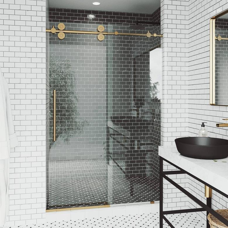 A frameless sliding shower door with gold matte finish and handle; a black subway shower wall and a white subway tile bathroom wall