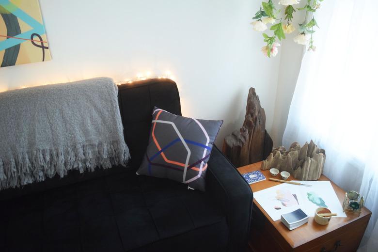 A corner of a room with a black couch near a window. There is art on the walls and a nearby chest with cypress wood knees surrounding it.