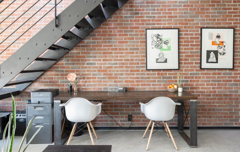 small spaces with Black metal staircase against brick wall with wood table, white curved chairs, framed print, and small metal cabinet