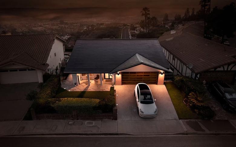 A ranch-style house at night with solar roof shingles; a Tesla is parked in front