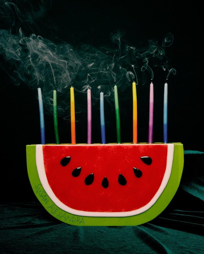 Watermelon-shaped menorah with colored candles on a black background.