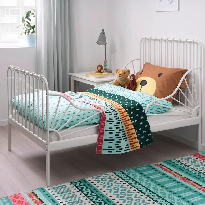 A white iron child's bed with a teddy bear pillow and colorful bedding