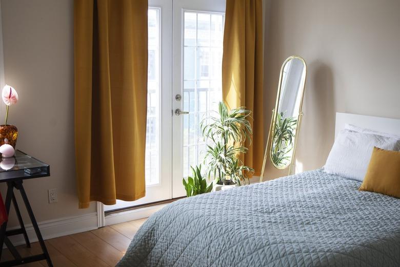 Bedroom with an oblong gold mirror, yellow curtains, and a desk with a pink lily flower. Bed with a blue blanket and yellow accent pillow.