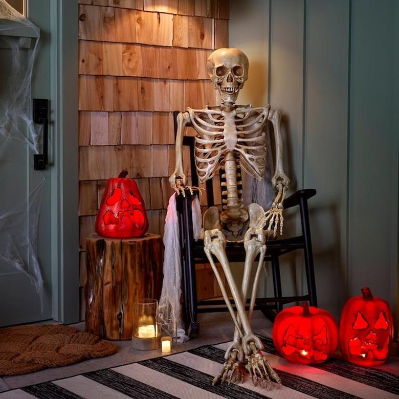 A fake skeleton sitting on a dark wood rocking chair on a front porch with light-up bright orange fake jack-'o-lanterns.