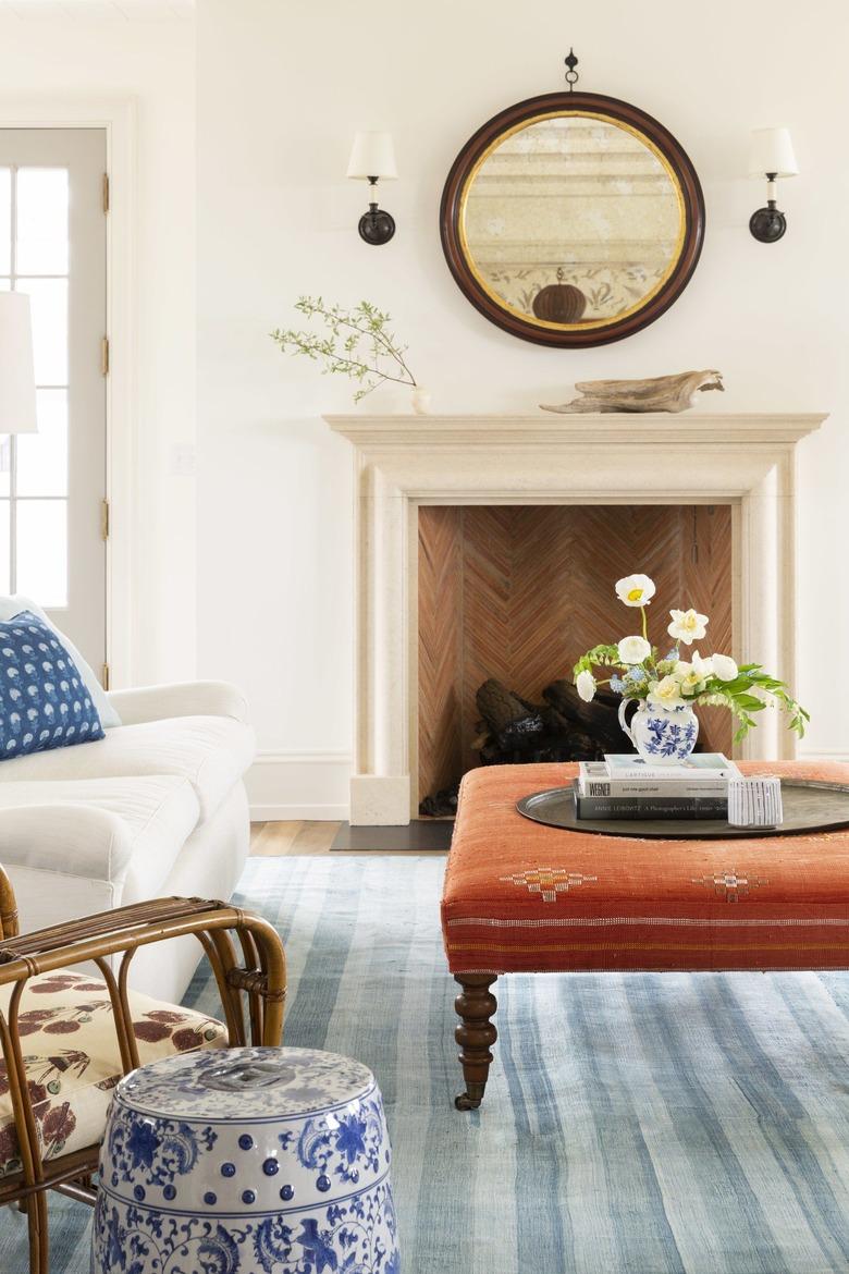 white living room with blue rug, orange ottoman and traditional fireplace with mirror on top