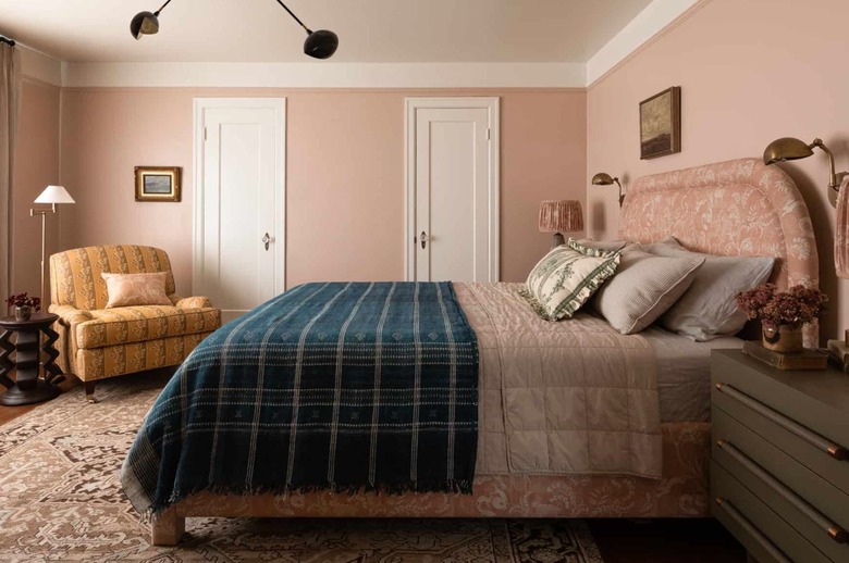 Bedroom with upholstered headboard, blanket, accent chair, end table, reading lamp, modern chandelier, pillows, sconces, nightstand, rug.