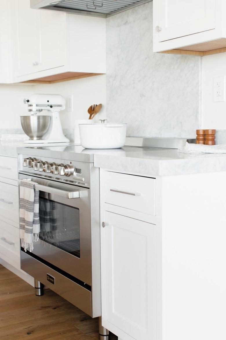 Kitchen counter with white drawers and a white marble countertop. In the middle of the counter, a stainless steel range. A white and grey striped kitchen towel is hanging over the handle over the oven door. A white dutch oven in on the stovetop, and there's a white KitchenAid stand mixer.