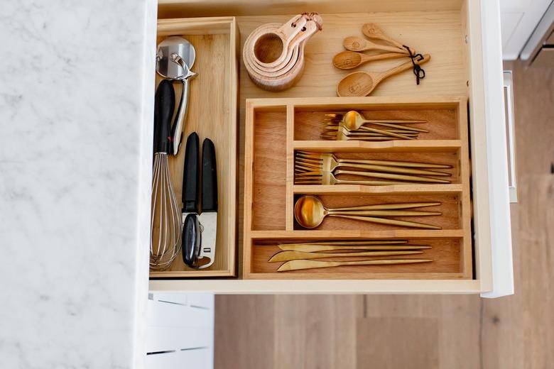 An open drawer with a wooden cutlery organizer. Gold cutlery, measuring cups and spoons, and other utensils visible inside. Marble countertop above the drawer.